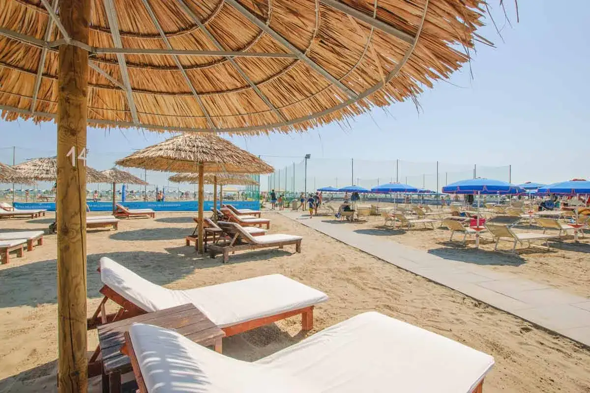 Spiaggia attrezzata con lettini e ombrelloni di paglia e blu, un percorso pedonale e campi da gioco sullo sfondo. Un perfetto bagno isola Cervia.