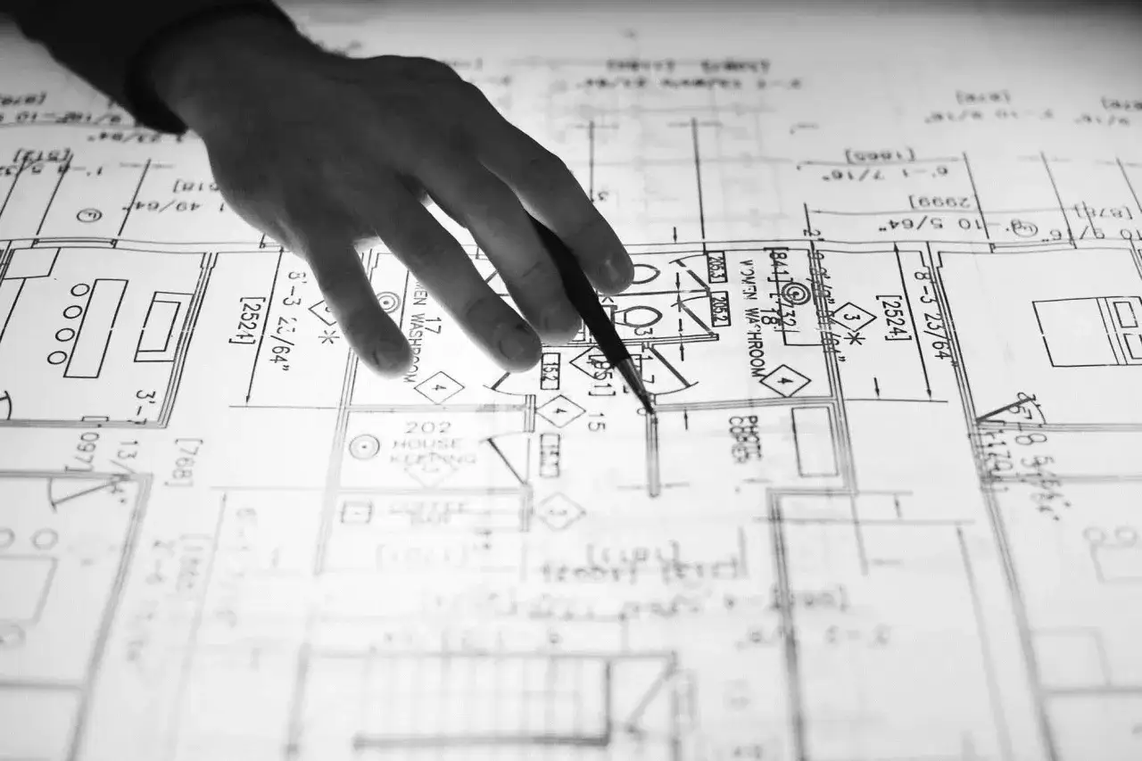 Mano di un disegnatore CAD che indica un progetto architettonico dettagliato, con misure e stanze definite.