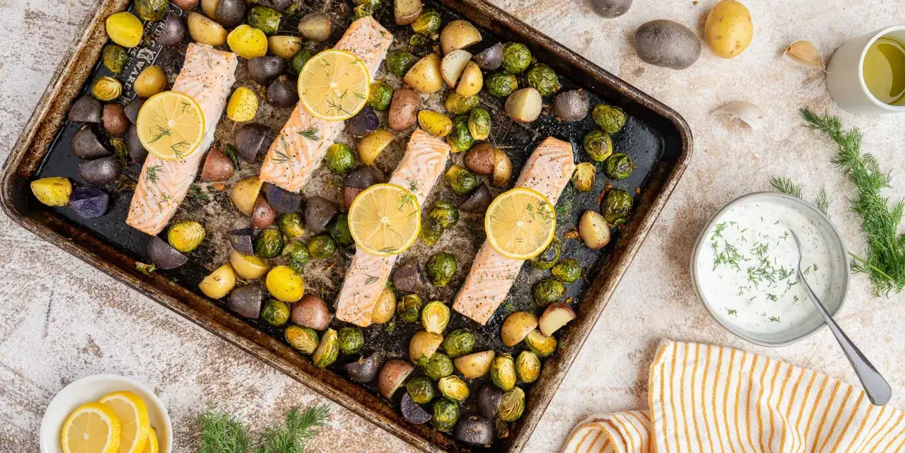 Quatre filets de saumon au four, garnis de rondelles de citron et d'aneth, accompagnés de pommes de terre rôties et de choux de Bruxelles sur une plaque.