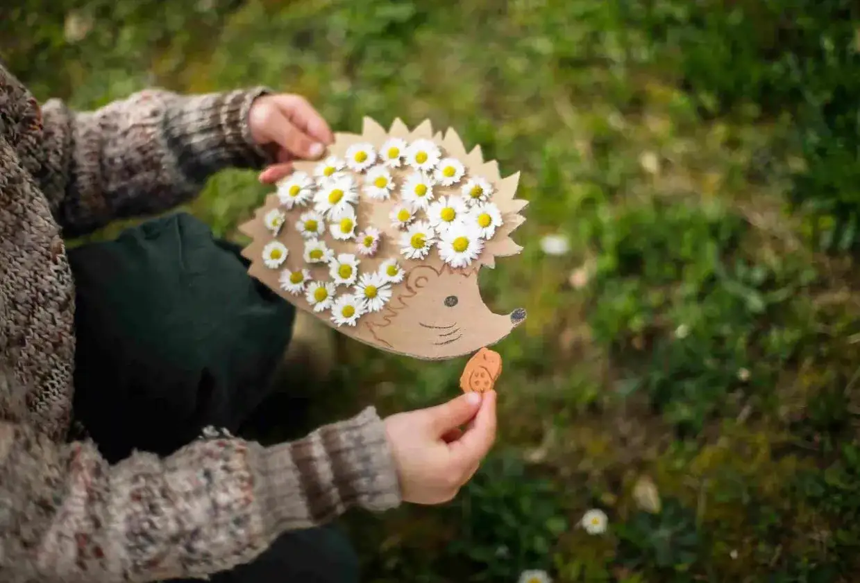 Ein Kind hält einen Igel aus Pappe, verziert mit Gänseblümchen. Ein tolles Bastelprojekt für den Kindergarten!