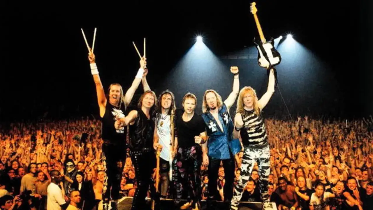 Les membres de Iron Maiden célèbrent sur scène devant une foule en délire, baguettes et guitare levées.