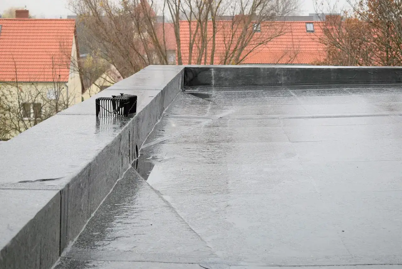 Mokry dach pokryty czarną membraną EPDM, z widocznym odpływem i budynkami w tle.