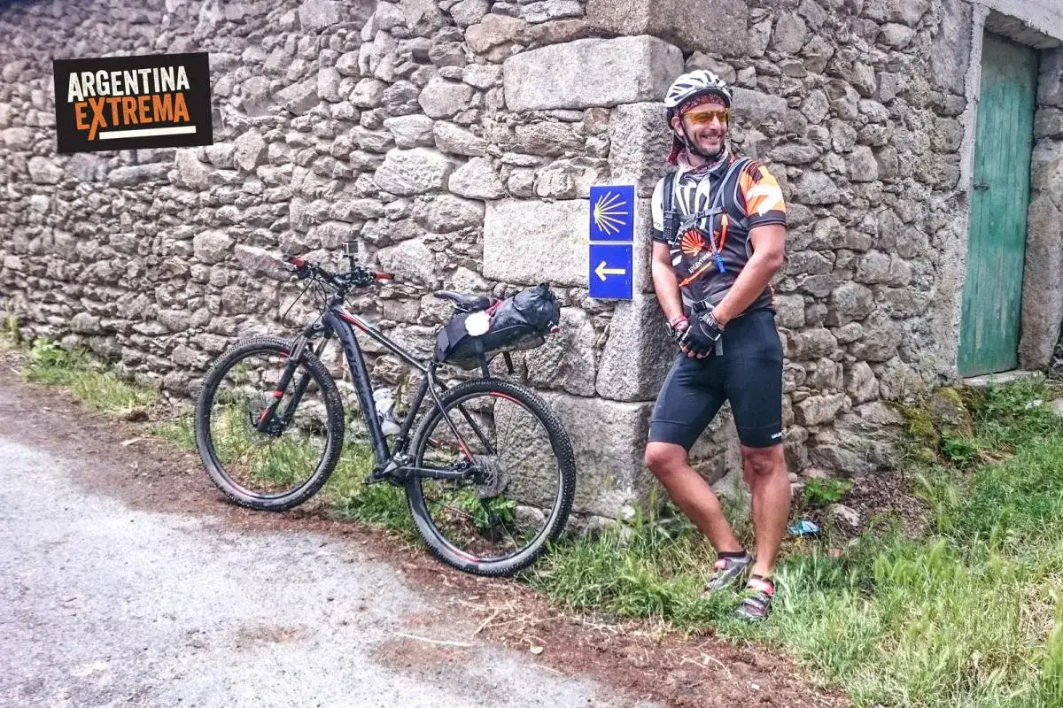 Ciclista sonriente junto a su bici, listo para el camino primitivo. Señal de flecha azul indica la ruta.