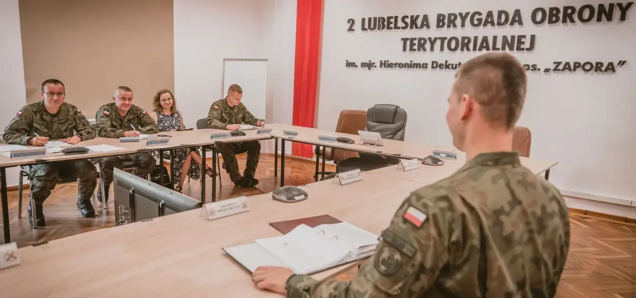 Żołnierze w mundurach siedzą przy stole w sali odpraw. Na ścianie widnieje napis 