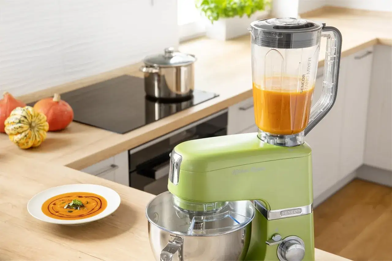 Zielony robot kuchenny z kielichem blendera pełnym zupy dyniowej. Na blacie stoją dynie i talerz z kremem. Idealny do tworzenia własnego **blender ręczny ranking**.