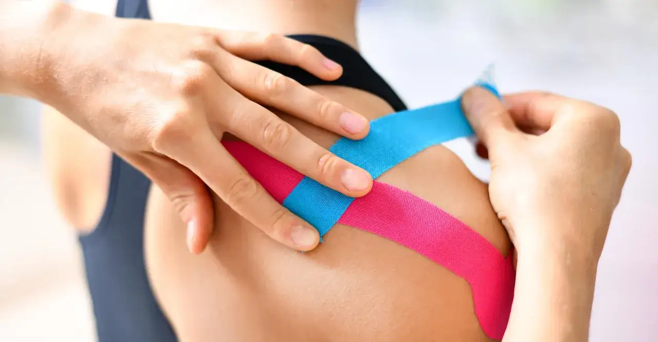 Hände legen pinkes und blaues Tape auf eine Schulter. Ein modernes Mittel gegen Schulterschmerzen, das an Omas Hausmittel erinnert.