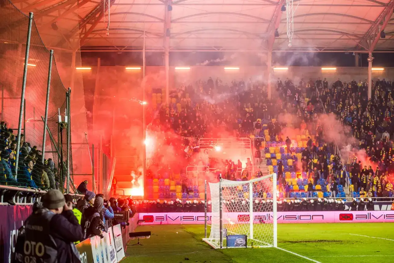 Gorąca atmosfera na stadionie podczas meczu. Kibice odpalają race, tworząc czerwony dym. To właśnie takie emocje towarzyszą prawdziwemu derby!