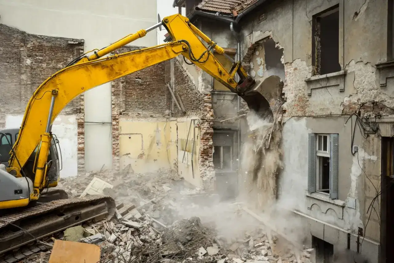 Żółta koparka prowadzi rozbiórkę starego budynku, wzbijając tumany kurzu.