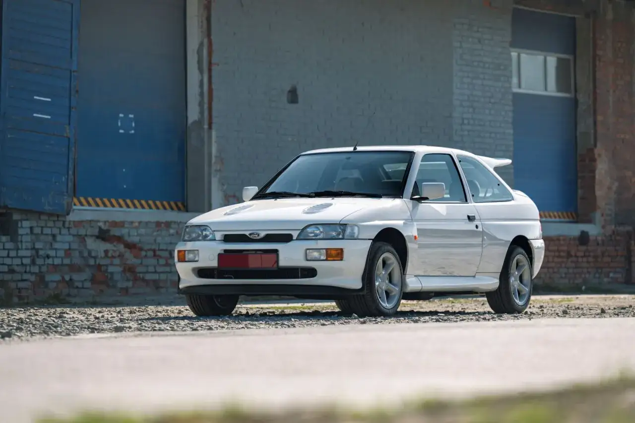 Biały Ford Escort Cosworth z charakterystycznym spojlerem i wlotami na masce, zaparkowany przed industrialnym budynkiem.