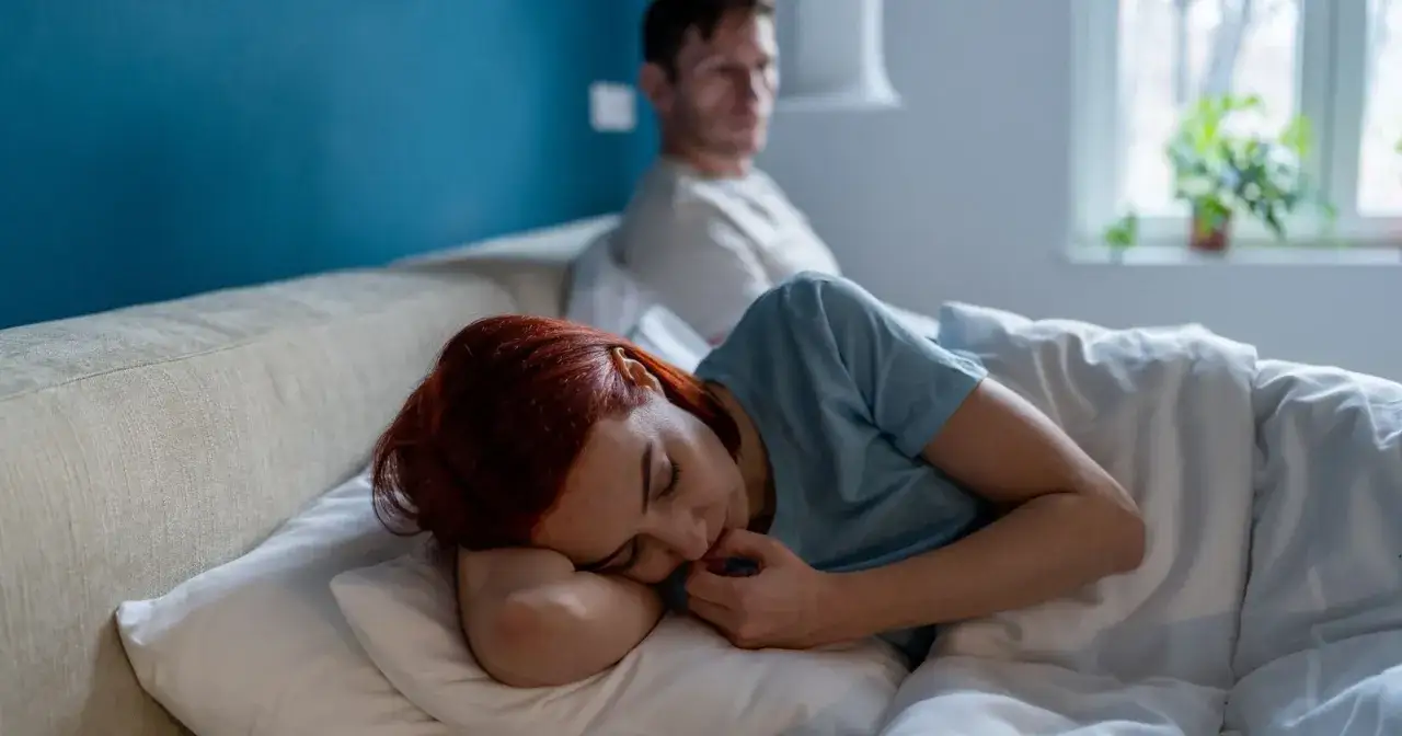Kobieta z rudymi włosami śpi, przytulając poduszkę. Mężczyzna siedzi z tyłu, odwrócony. Czuje się nieważna dla partnera.