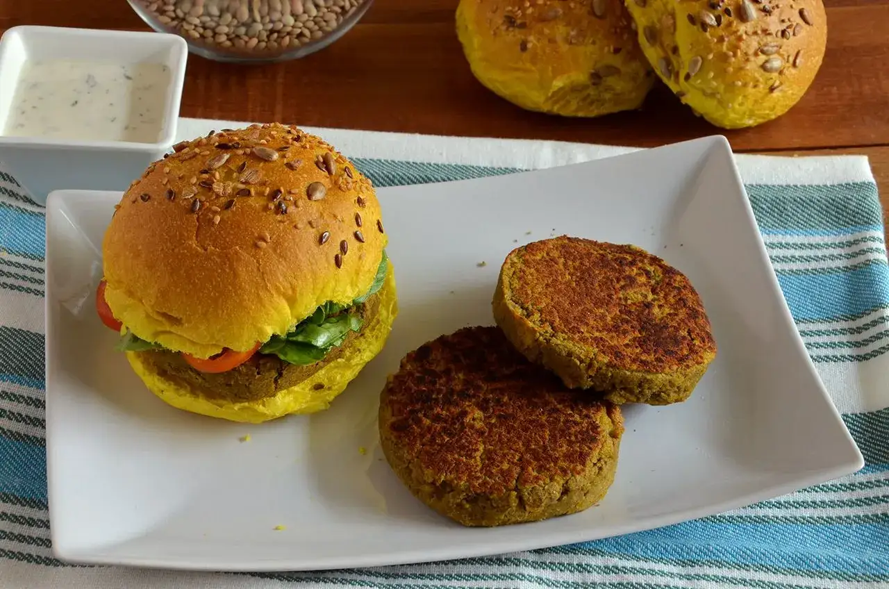 Hamburger di lenticchie dorato, con semi, lattuga e pomodoro. Due polpette pronte da gustare accanto.