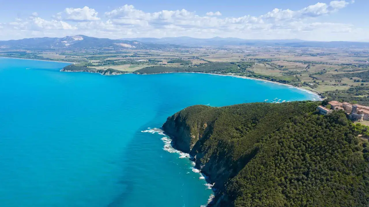 Vista aerea della costa degli etruschi con mare turchese, scogliere verdi e un borgo storico.