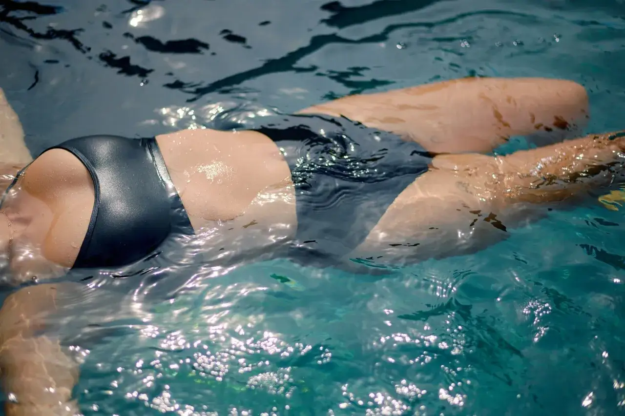 A person floats in a pool, wearing a dark swimsuit. The question