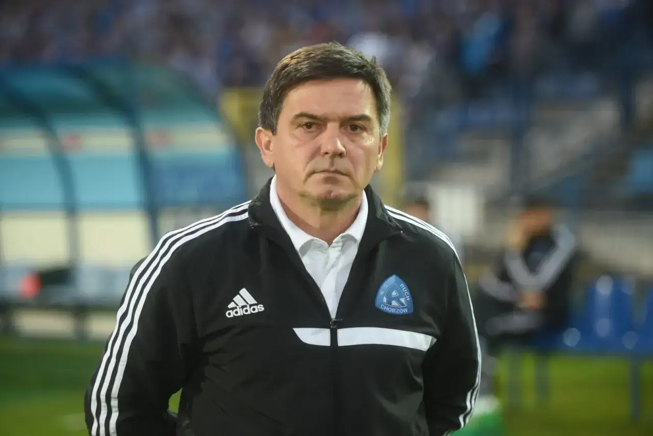 Poważny trener Ruchu Chorzów w czarnej kurtce z logo klubu i Adidasa.