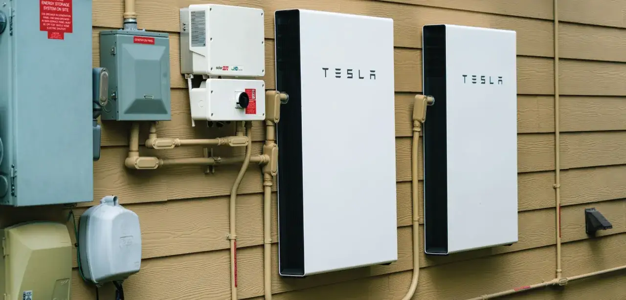 Dwa białe magazyny energii elektrycznej Tesla zamontowane na brązowej ścianie domu, obok paneli elektrycznych i okablowania.