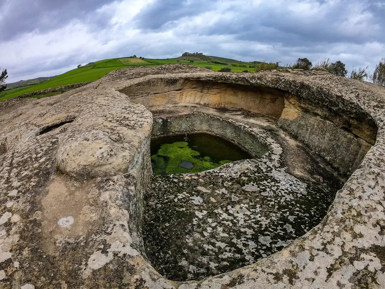 Antica domus de janas s'incantu, scavata nella roccia, con vasca d'acqua verde e colline sullo sfondo.