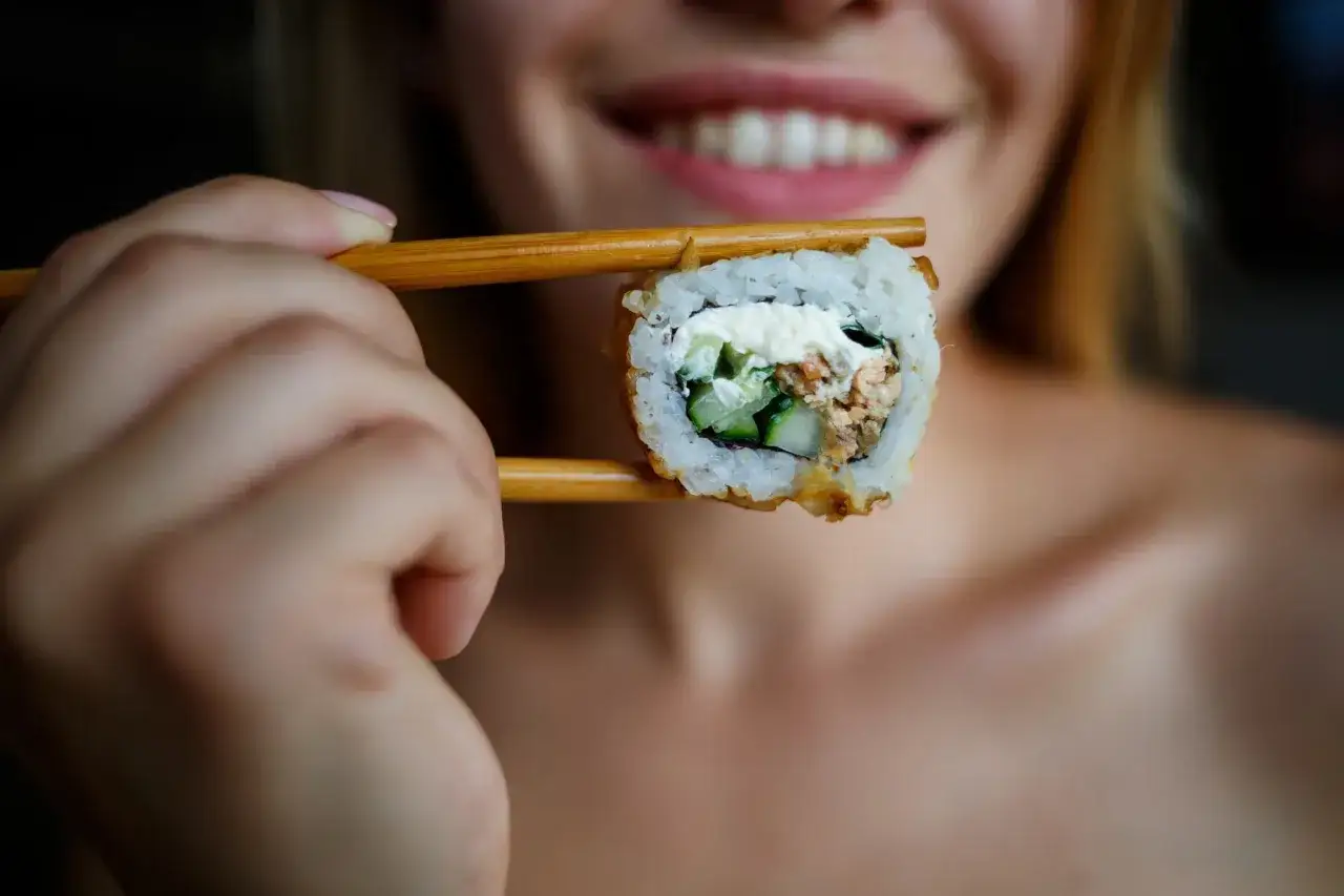 Uśmiechnięta kobieta trzyma pałeczkami sushi z łososiem i og&oacute;rkiem. Czy można jeść sushi karmiąc piersią?
