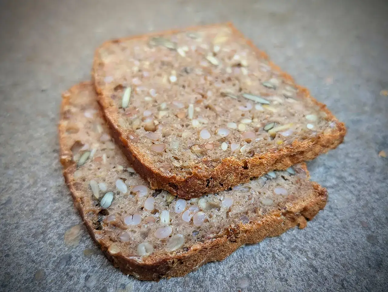 Zwei Scheiben Vollkornbrot mit vielen K&ouml;rnern, darunter auch Weizenkorn, liegen auf grauem Untergrund.