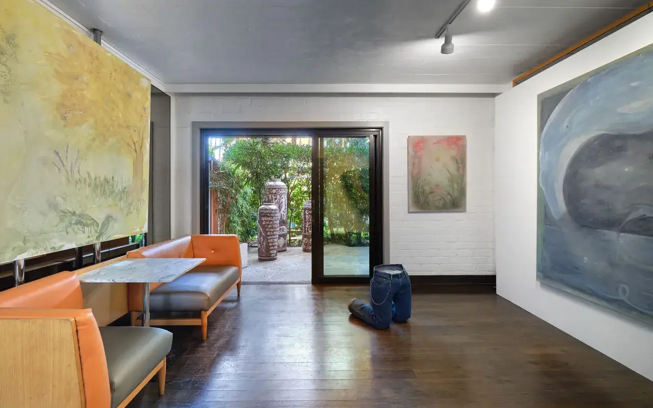 A contemporary space showcasing young art galleries, featuring abstract paintings, a surreal denim sculpture, and an outdoor patio visible through glass doors.