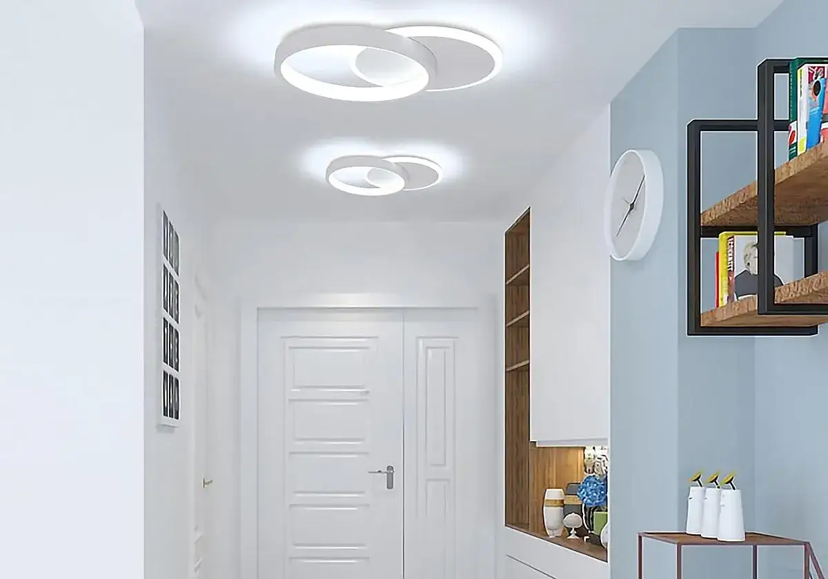 Dos modernos plafones cuadrados de techo iluminan un pasillo con puertas blancas y estanterías.
