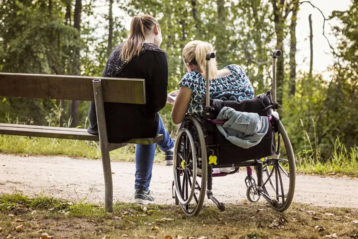 Opiekun osoby niepełnosprawnej siedzi na ławce obok kobiety na wózku inwalidzkim w parku.
