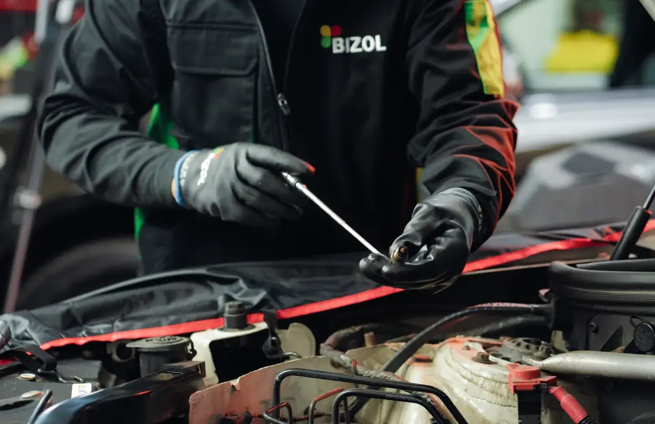Mechanik w czarnej kurtce z logo BIZOL sprawdza świecę zapłonową, być może szukając przyczyny stukania na zimnym silniku diesel.