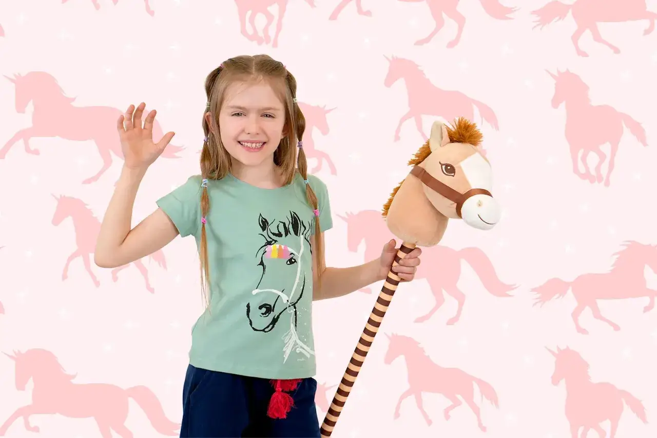Uśmiechnięta dziewczynka z konikiem na patyku, gotowa do zabawy w hobby horsing na tle r&oacute;żowych jednorożc&oacute;w.