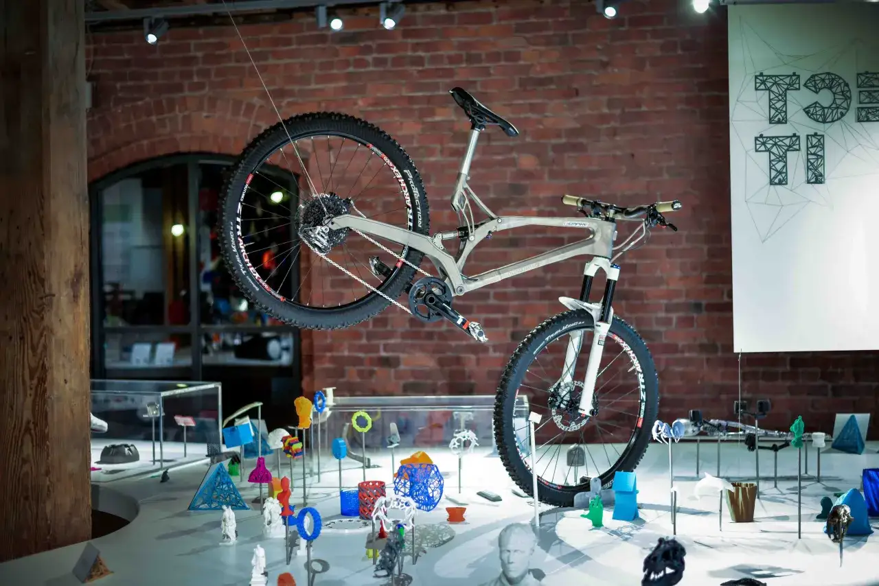 A 3d printed bike hangs suspended, showcasing innovative design. The frame's intricate lattice structure is visible, surrounded by various 3D printed objects.