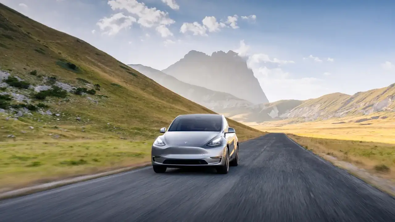 Srebrny Tesla Y jedzie pustą drogą przez górzysty krajobraz. Imponujący zasięg pozwala na odkrywanie dzikich zakątków.