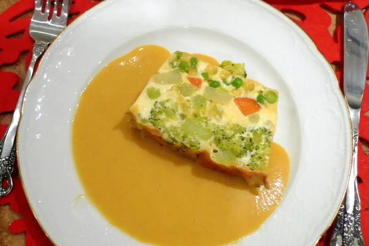 Porción de pastel de verduras con brócoli, zanahoria y guisantes, servida con una cremosa salsa.