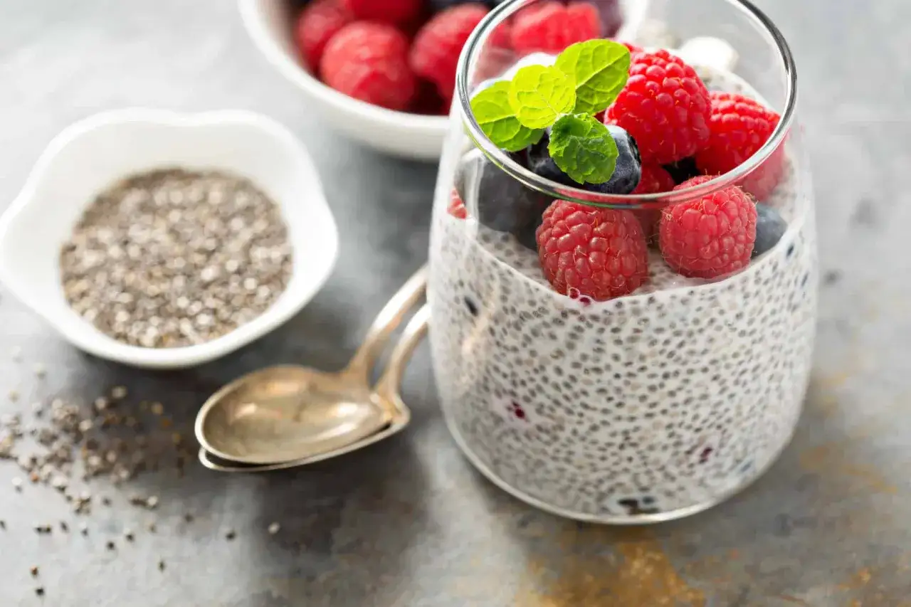 Pudding z nasion chia z malinami i borówkami, ozdobiony miętą. Obok miseczka z nasionami chia i łyżeczka.