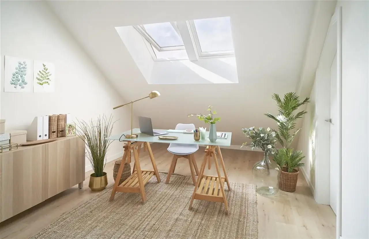 Un estudio luminoso bajo **ventanas abuhardilladas**, con escritorio, plantas y un mueble de madera.