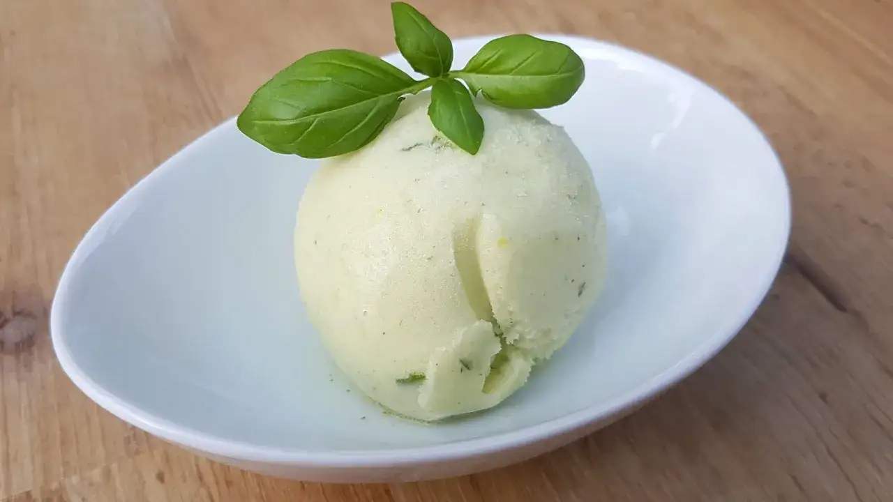 Ein Löffel Basilikum Eis, garniert mit frischen Basilikumblättern, serviert in einer weißen Schale auf Holzhintergrund.