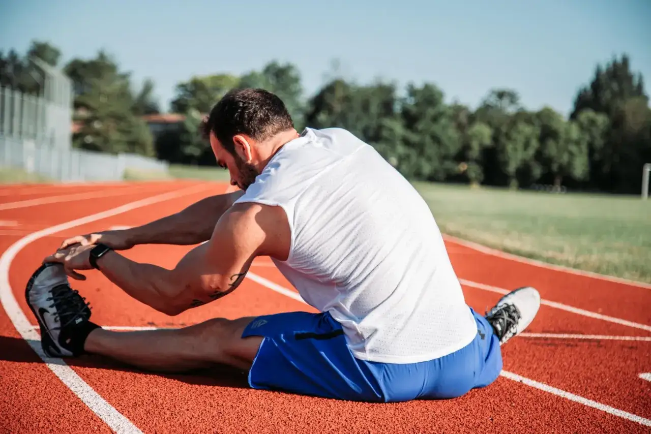 Mężczyzna w białej koszulce i niebieskich spodenkach wykonuje **stretching** na bieżni.