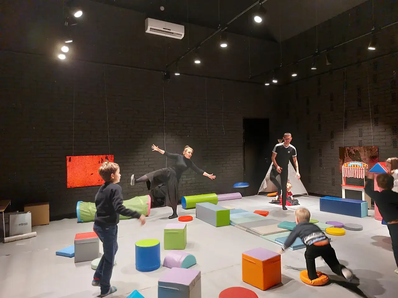 Dzieci i dorośli bawią się w sali gimnastycznej, która służy jako teatr dla młodzieży. Kolorowe klocki i materace tworzą scenografię.