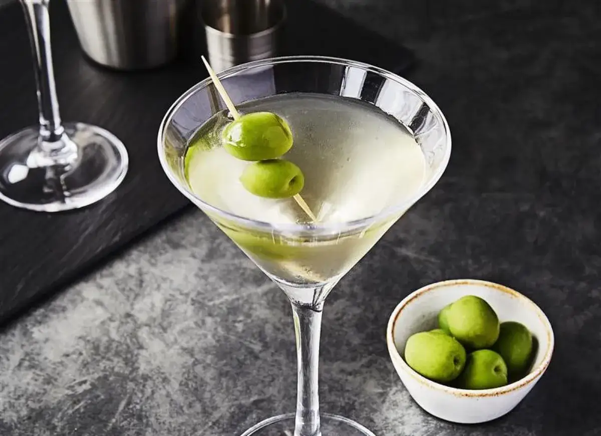 Elegancki martini z wódką, ozdobione dwoma oliwkami na wykałaczce. Obok miseczka z kilkoma zielonymi oliwkami.