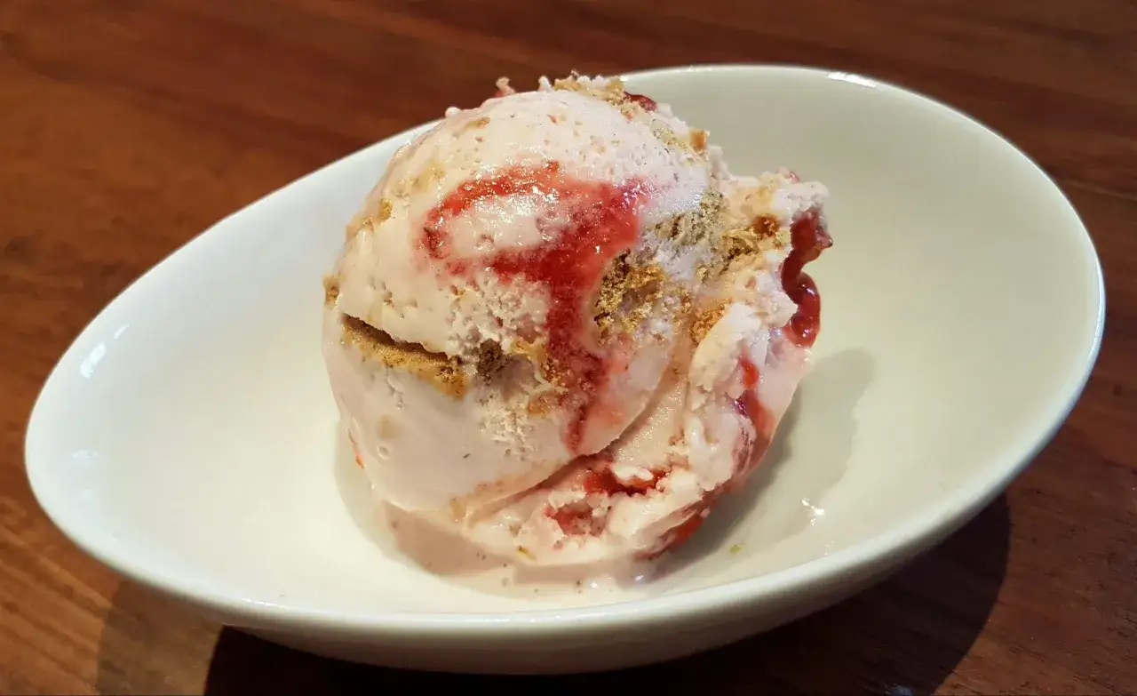 Ein Löffel cremiges strawberry cheesecake eis mit Erdbeersauce und Keksstückchen in einer weißen Schale.