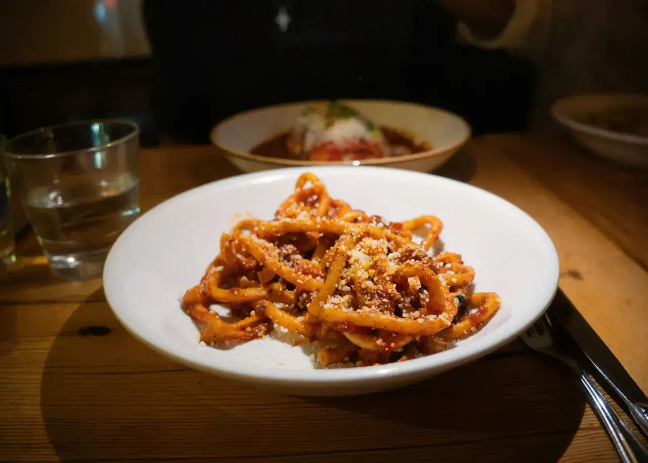 Herzhaftes Amatriciana Rezept auf einem Teller, garniert mit geriebenem K&auml;se. Daneben ein Glas Wasser und Besteck.