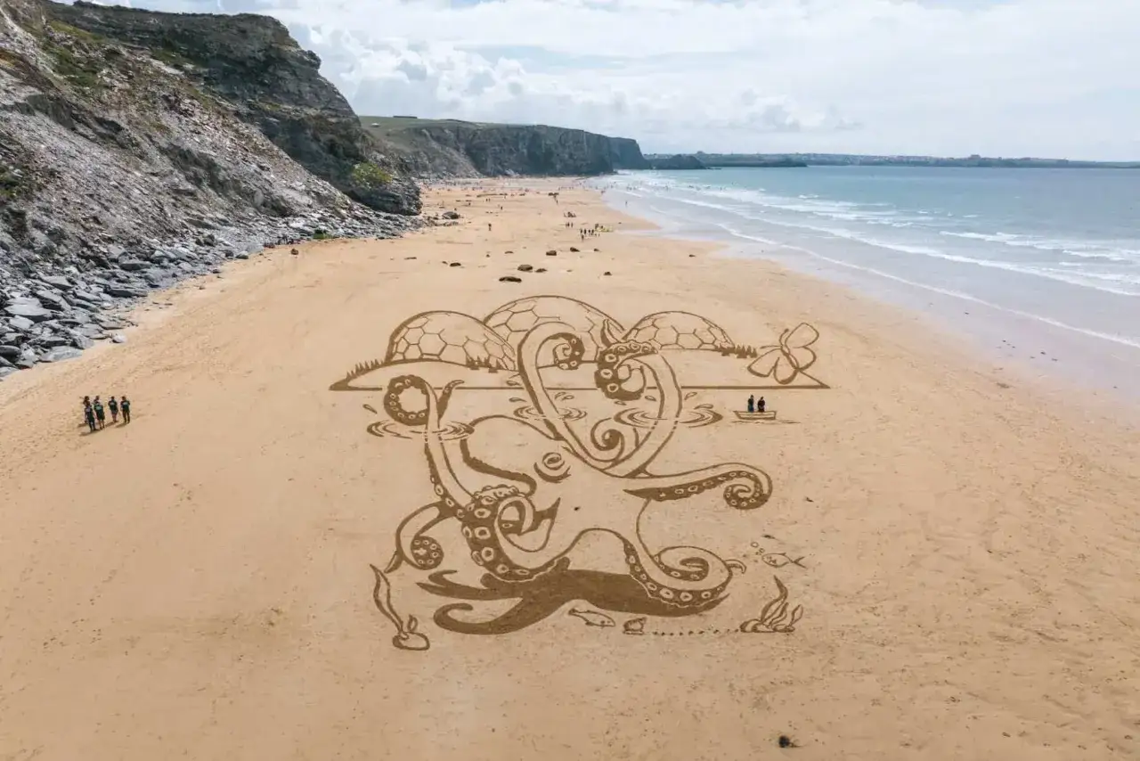 Enorm bläckfisksandskulptur på stranden i St Ives, med klippor och hav i bakgrunden.