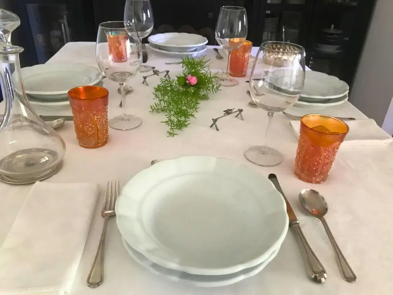 Tavola apparecchiata con cura, bicchieri di cristallo, piatti bianchi e posate d'argento. Un tocco di verde e un fiorellino rosa al centro, per un'eleganza che rispetta il galateo.
