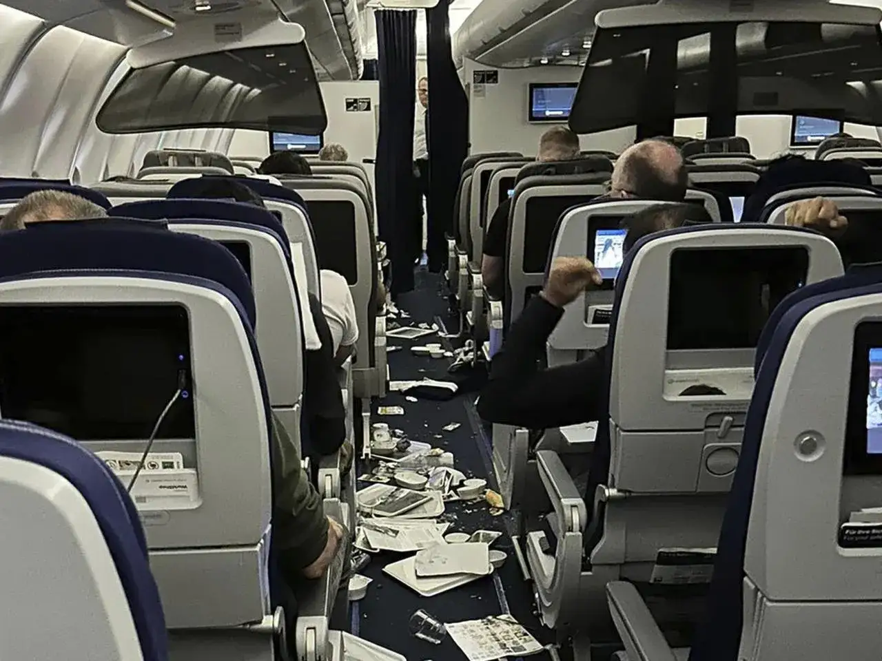Wnętrze samolotu po silnych turbulencjach. Na podłodze leżą porozrzucane przedmioty, kubki i gazety. Pasażerowie siedzą na swoich miejscach.