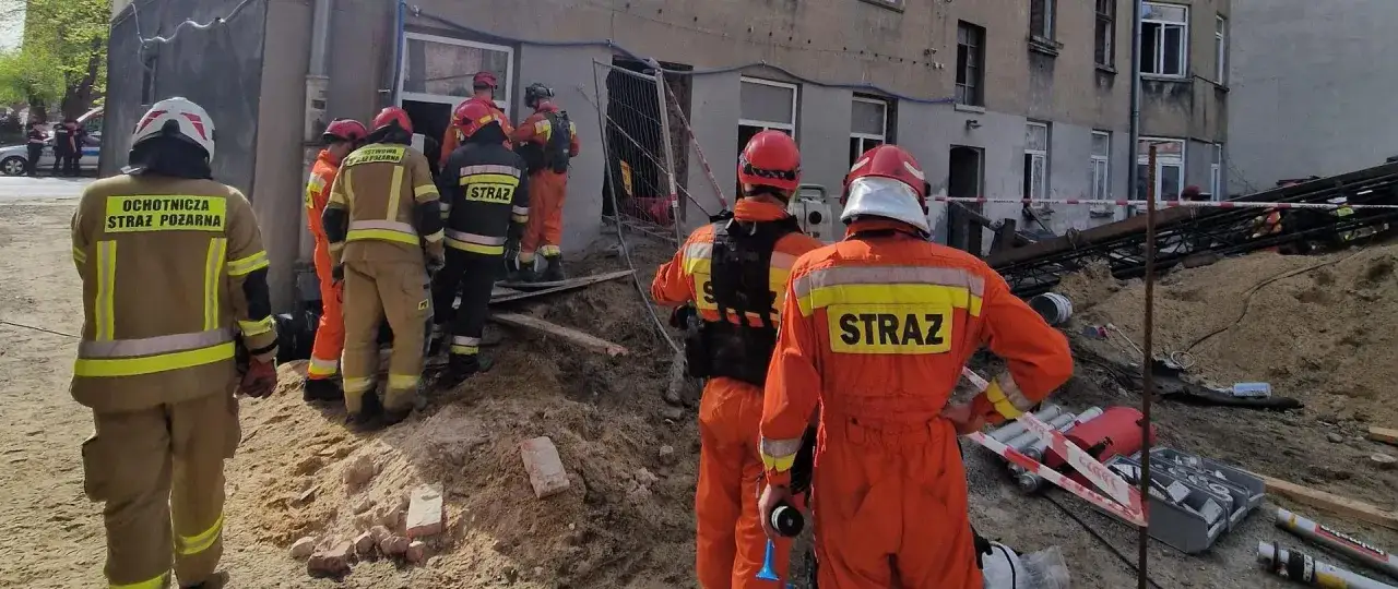 Strażacy w akcji po zawaleniu się budynku. Grupa ratowników w pomarańczowych i beżowych kombinezonach zabezpiecza teren i ocenia szkody.
