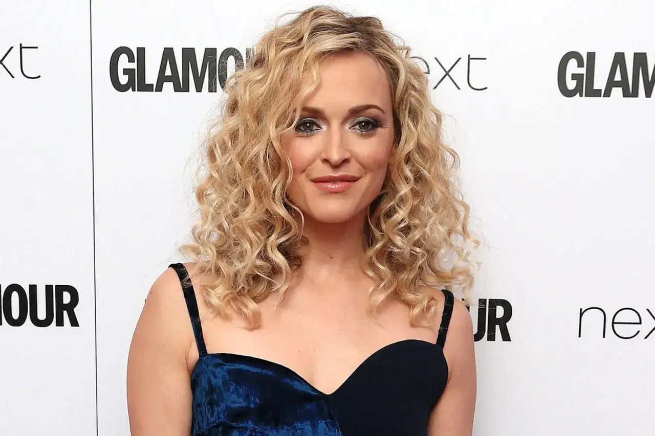 Une femme avec une coiffure ann&eacute;e 80, des boucles blondes volumineuses et un maquillage audacieux. Elle porte une robe bleue.