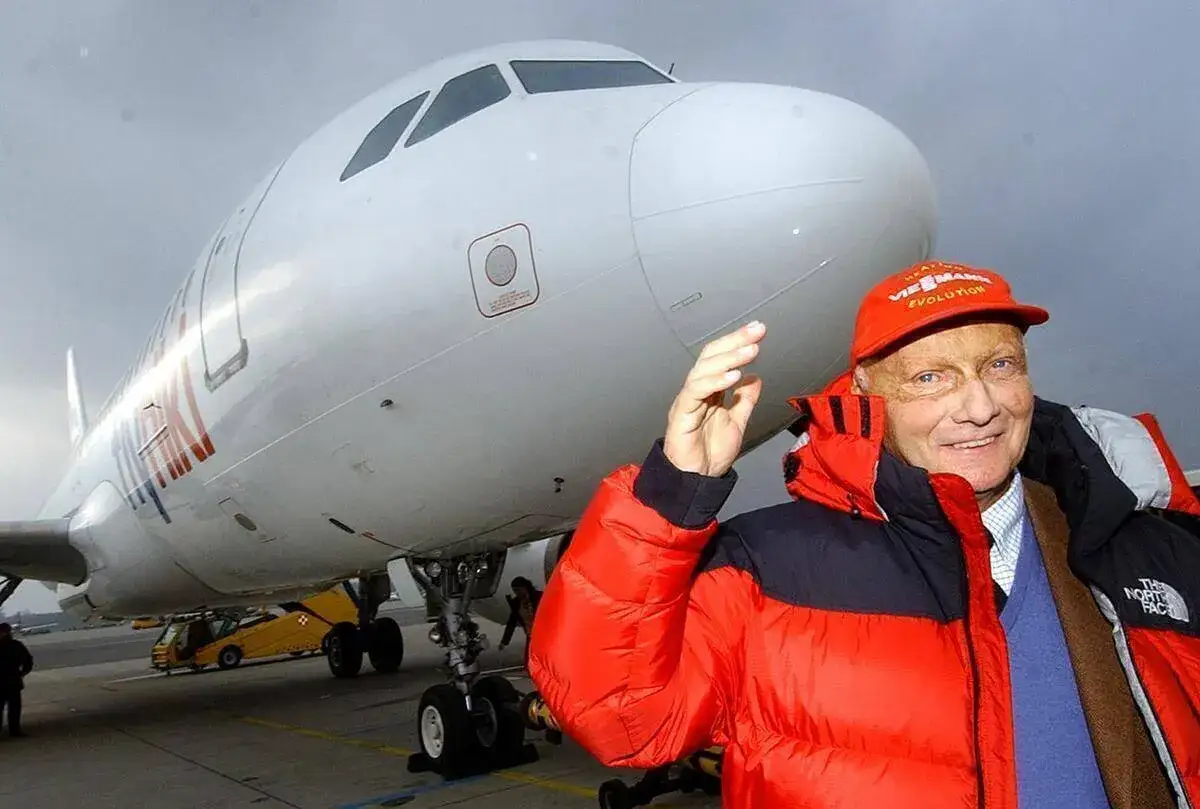 Niki Lauda, w czerwonej kurtce i czapce, macha ręką przed samolotem Lauda Air.