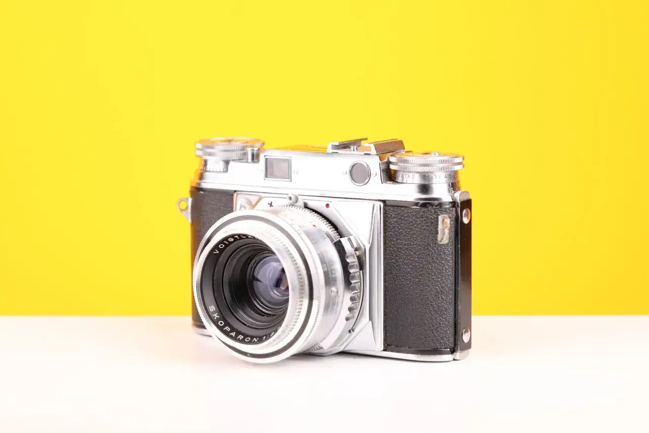 A vintage Minolta CL camera with a Voigtlander Skopar lens sits on a white surface against a bright yellow background.