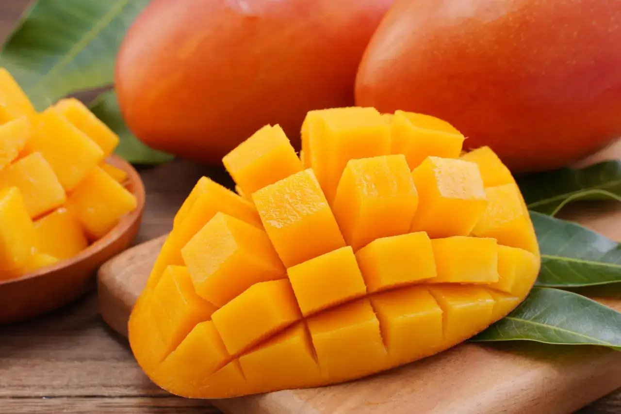 Dojrzałe mango, pokrojone w kostkę, prezentuje swoje soczyste, pomarańczowe wnętrze. Obok leżą całe owoce i liście.