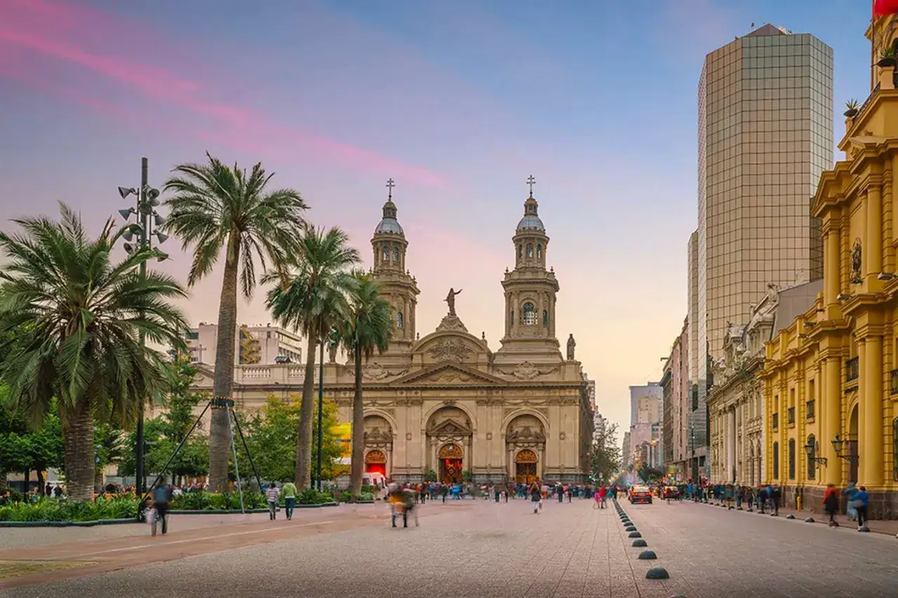 Zachód słońca nad placem w Santiago de Chile. Majestatyczna katedra z dwiema wieżami góruje nad palmami i nowoczesnymi budynkami.