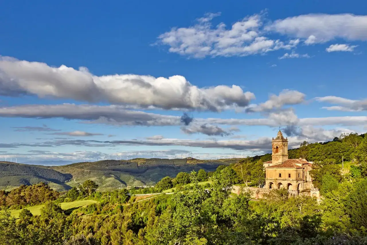 Iglesia con torre campanario en una colina verde, rodeada de árboles y con molinos de viento al fondo. Las torres del río vigilan el paisaje.