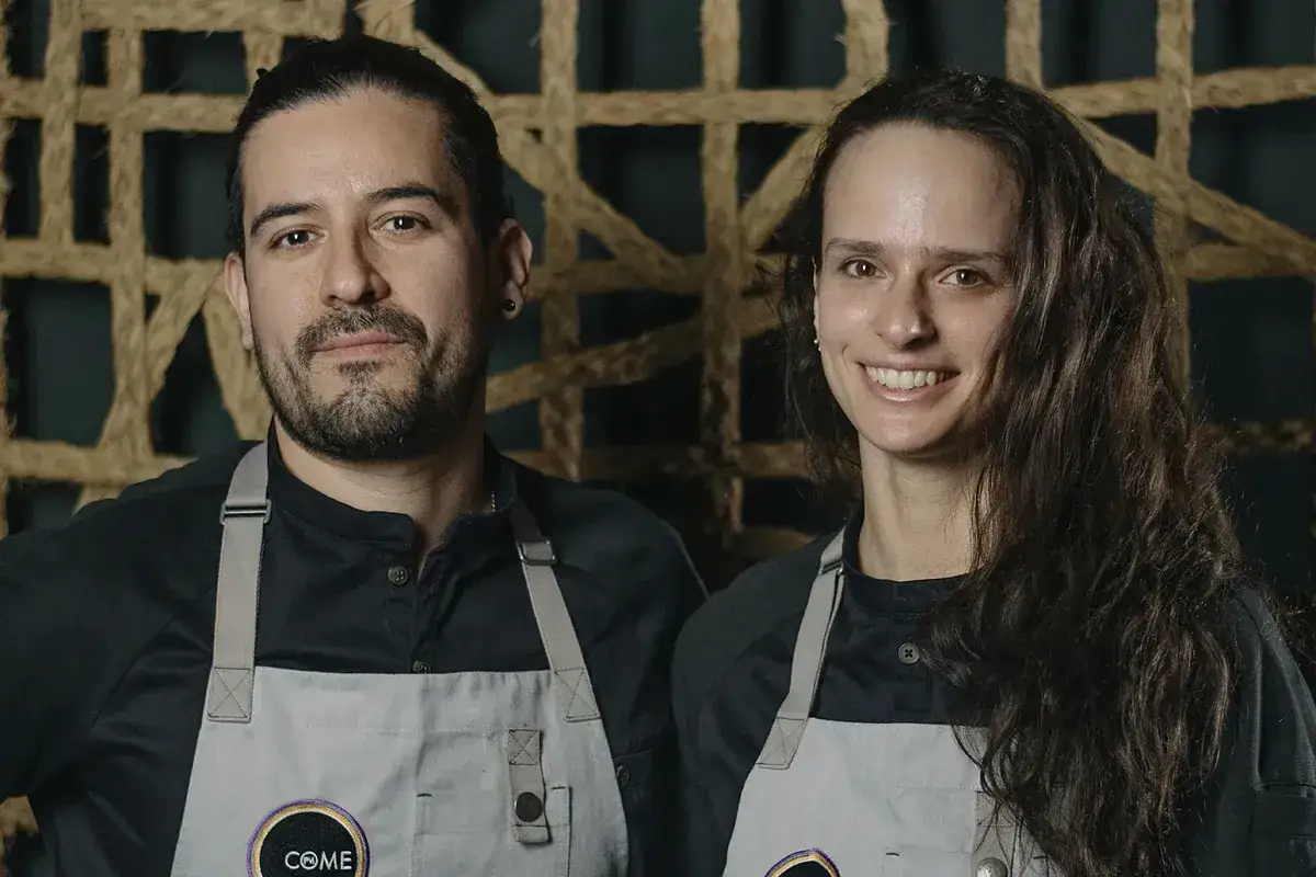 Dos chefs sonríen, uno con coleta y barba, la otra con cabello largo. Ambos llevan delantales con el logo