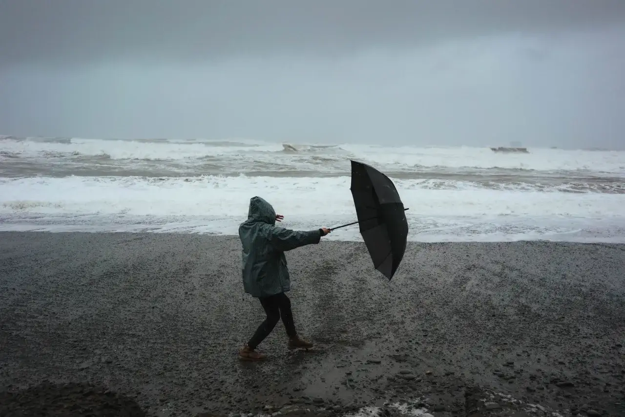 Osoba w deszczówce walczy z wiatrem i parasolem na plaży. W tle widać wzburzone morze, oddające aktualny stan pogody.